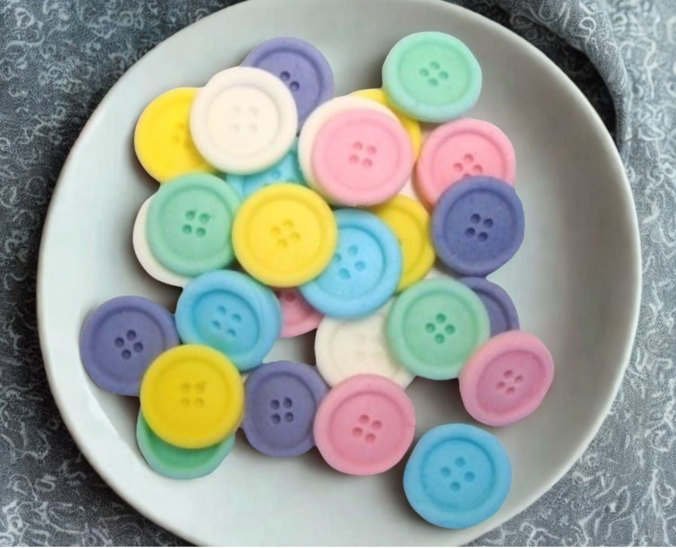 30 mixed colored edible buttons on a white plate against a textured gray background