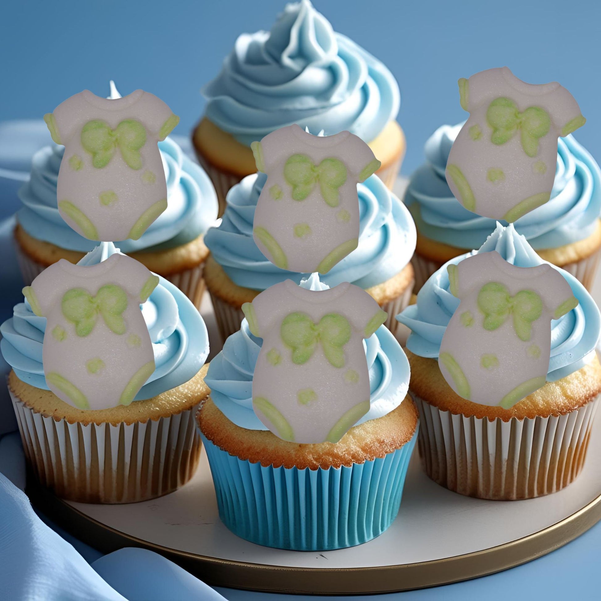 Cupcakes with blue frosting and edible green baby onesie decorations on a plate.