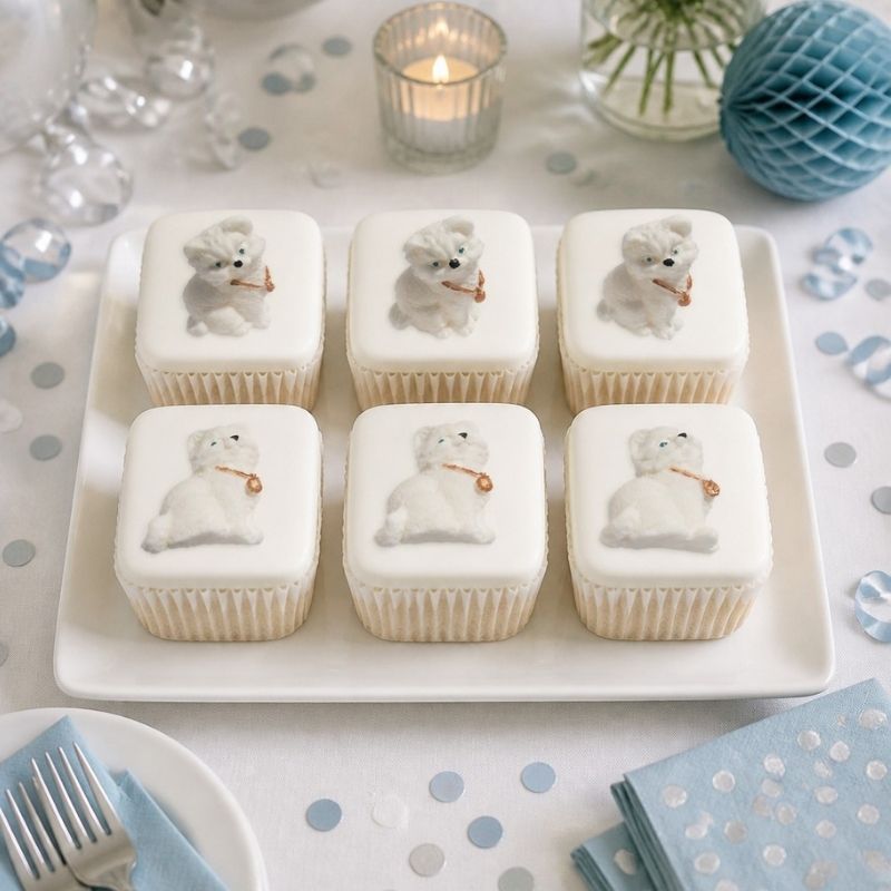 Six white-frosted cupcakes arranged on a white rectangular platter, each topped with a small white kitten cat . The cupcakes are styled on a decorated table with blue napkins, confetti, a glass candle holder, and blue paper decor.