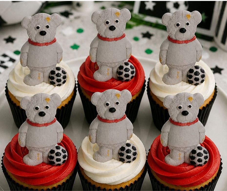 Six decorated cupcakes arranged in two rows, each topped with a small gray teddy bear figurine wearing a red collar and accompanied by a black-and-white football topper. The cupcakes are frosted in alternating white and red swirls, set on a white plate with a star-patterned background.