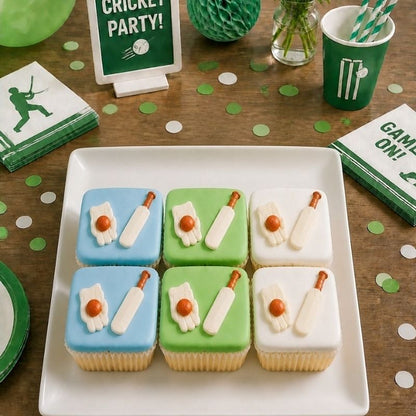 Six iced cupcakes arranged on a white square plate, decorated with light blue, light green, and white frosting, each with a white cricket bat and a small glove with orange ball detail. The cupcakes are photographed on a wooden table with cricket party decorations, including a green cup, napkins, and a sign reading 'Cricket Party!'