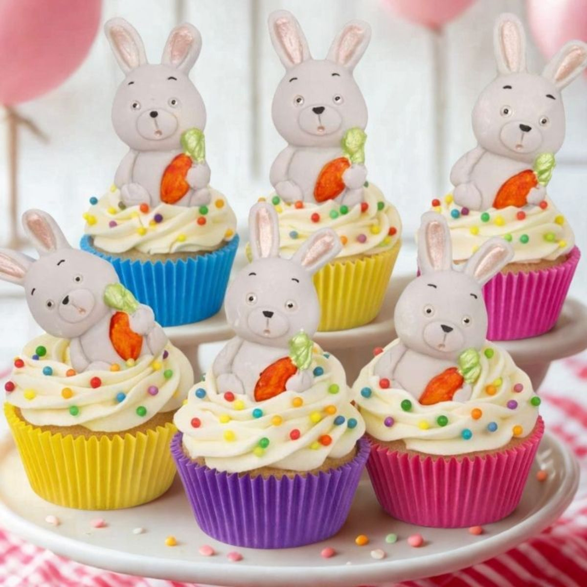 Six bunny-shaped cupcake toppers decorated with white frosting, colourful sprinkles, and orange carrots, displayed on a white cake stand.